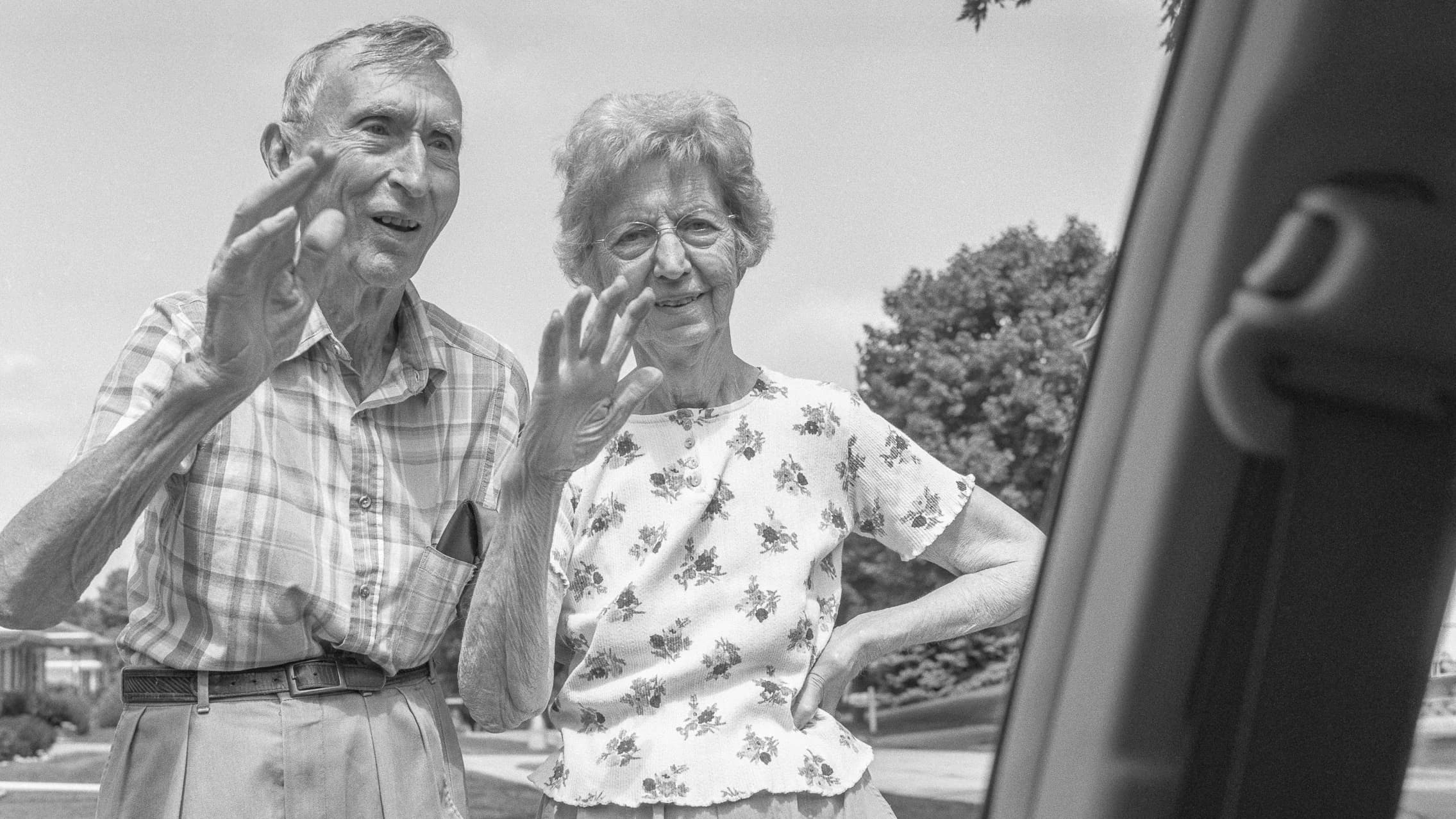 An elderly couple stand together outside, smiling and waving warmly